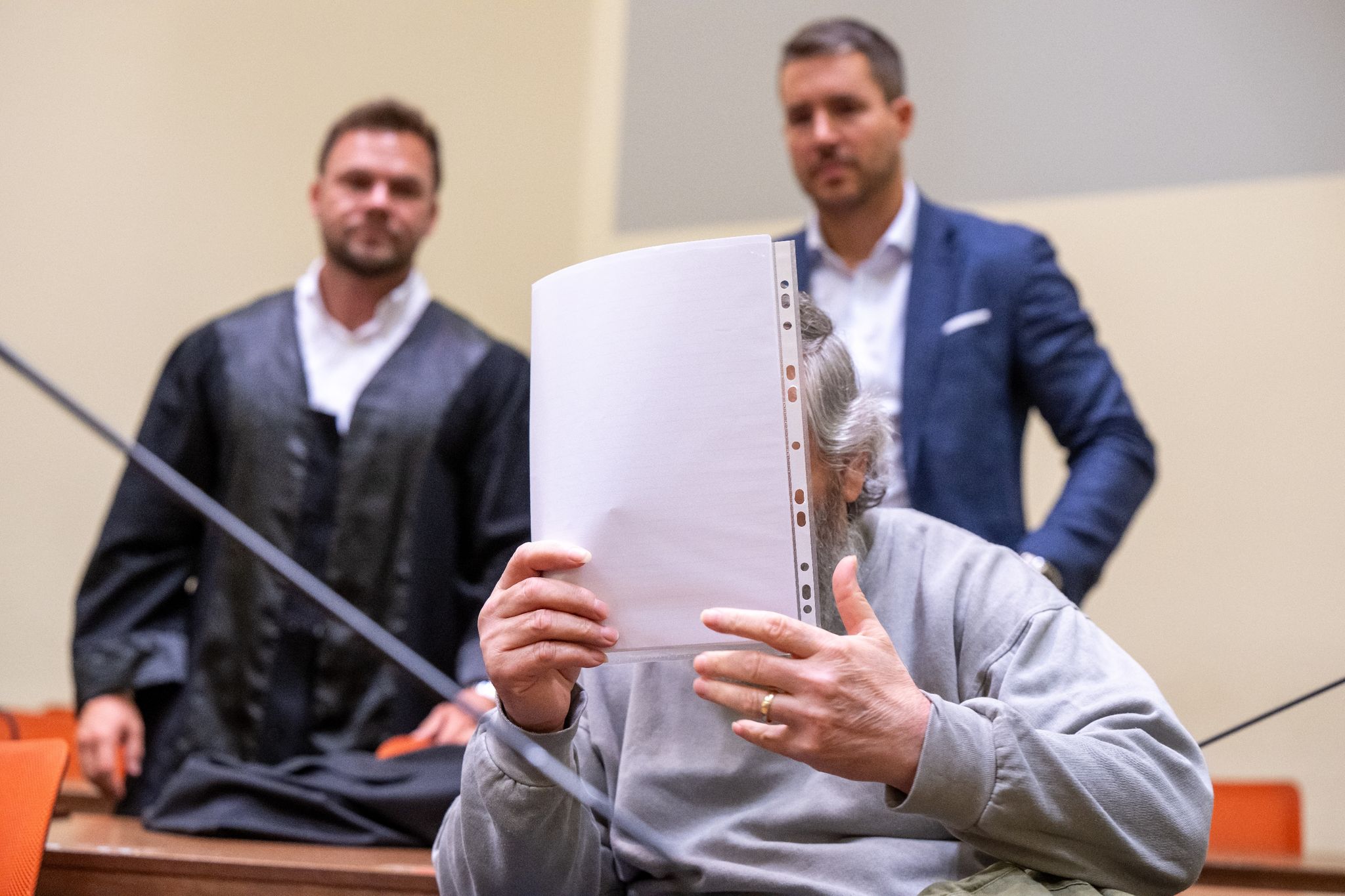Der Angeklagte hält sich im Gerichtssaal ein Blatt Papier vor das Gesicht. Im Hintergrund stehen seine Anwälte. (© Peter Kneffel/dpa)